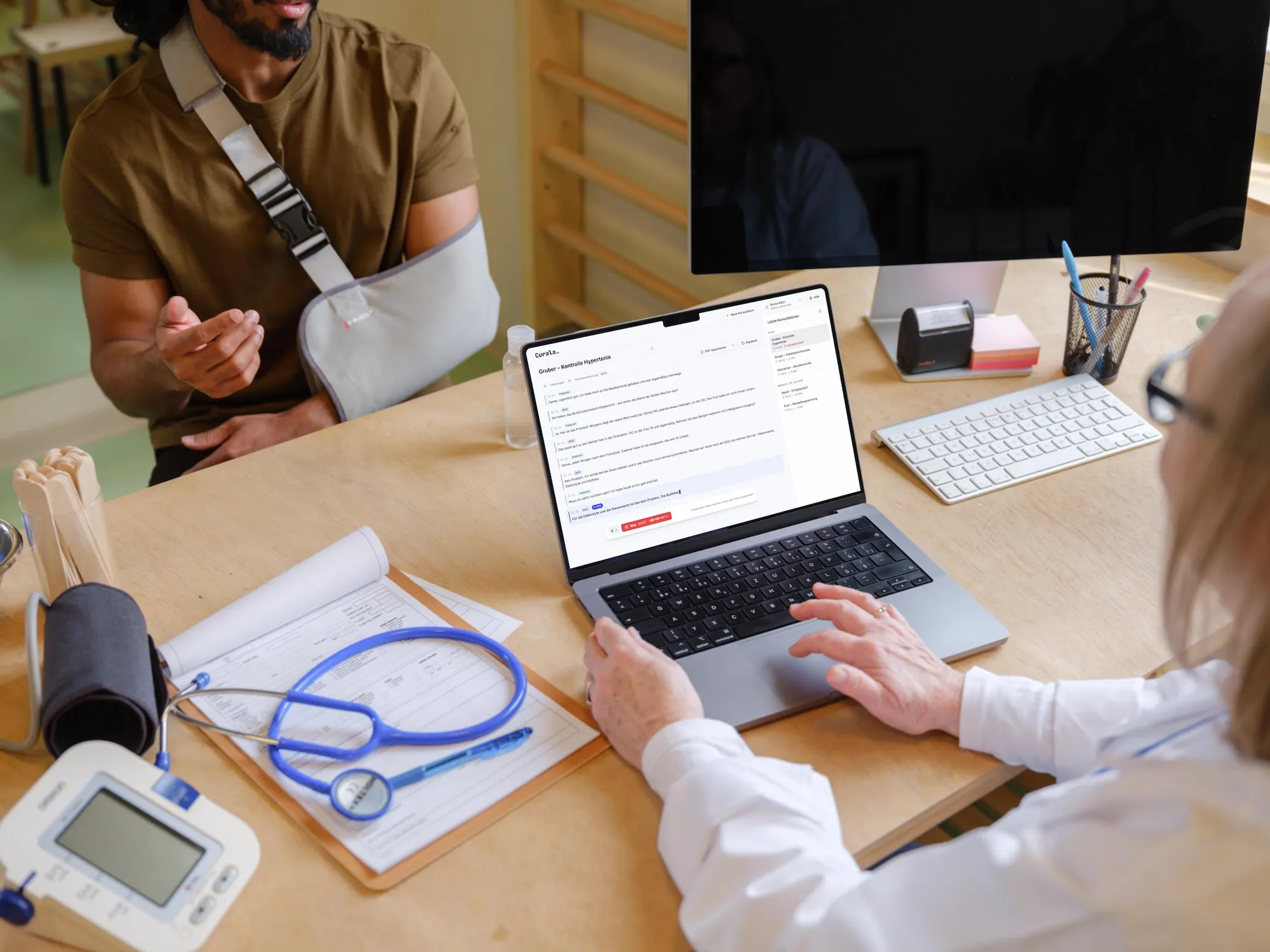 Ärztin im Gespräch mit einem Patienten, Curala transkribiert die Konsultation nebenher auf dem Laptop